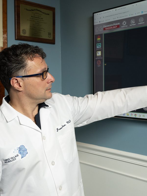 In his office adorned with framed certificates, a doctor from Three Village Neurology, wearing a white coat, points to a brain scan on the screen, exemplifying the clinic's commitment to exceptional neurological care.