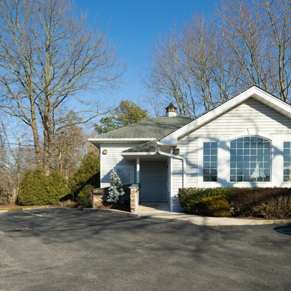 Single-story building with gray siding, large windows, and a signboard on the left that reads "Three Village Neurology." Surrounded by trees, this neurology clinic features an asphalt parking area. Single-story building with gray siding, large windows, and a signboard on the left that reads "Three Village Neurology." Surrounded by trees, this neurology clinic features an asphalt parking area.